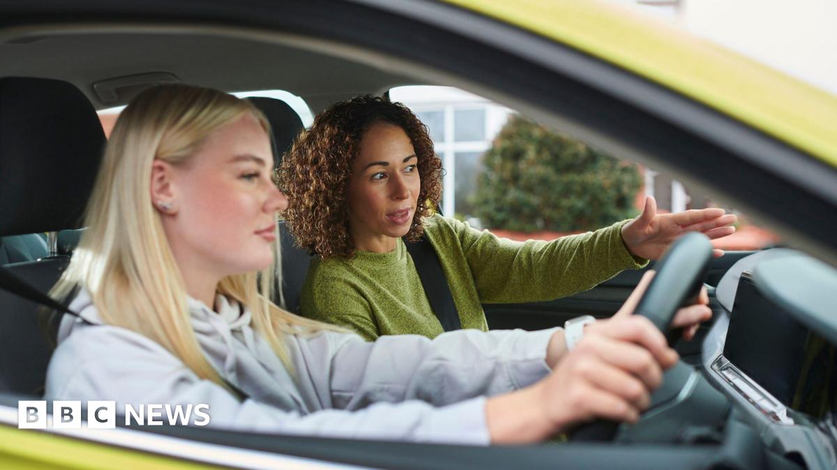 Two women in a car. The younger one is at the wheel. She is white and has blonde hair and is wearing a hoodie. An older mixed-race woman is instructing her. The car is yellow-green