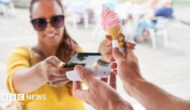 A woman dressed in a yellow top and wearing sunglasses buys a pink ice cream by placing her smartphone near a payment terminal.