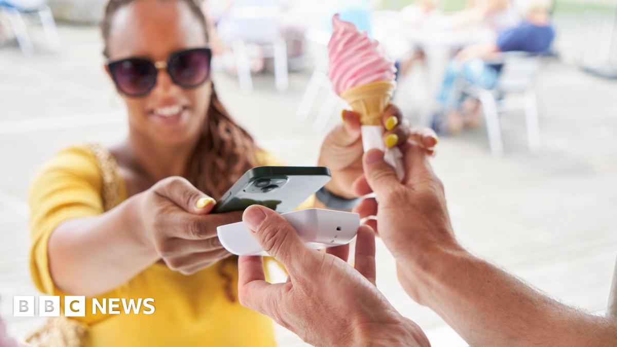 A woman dressed in a yellow top and wearing sunglasses buys a pink ice cream by placing her smartphone near a payment terminal.
