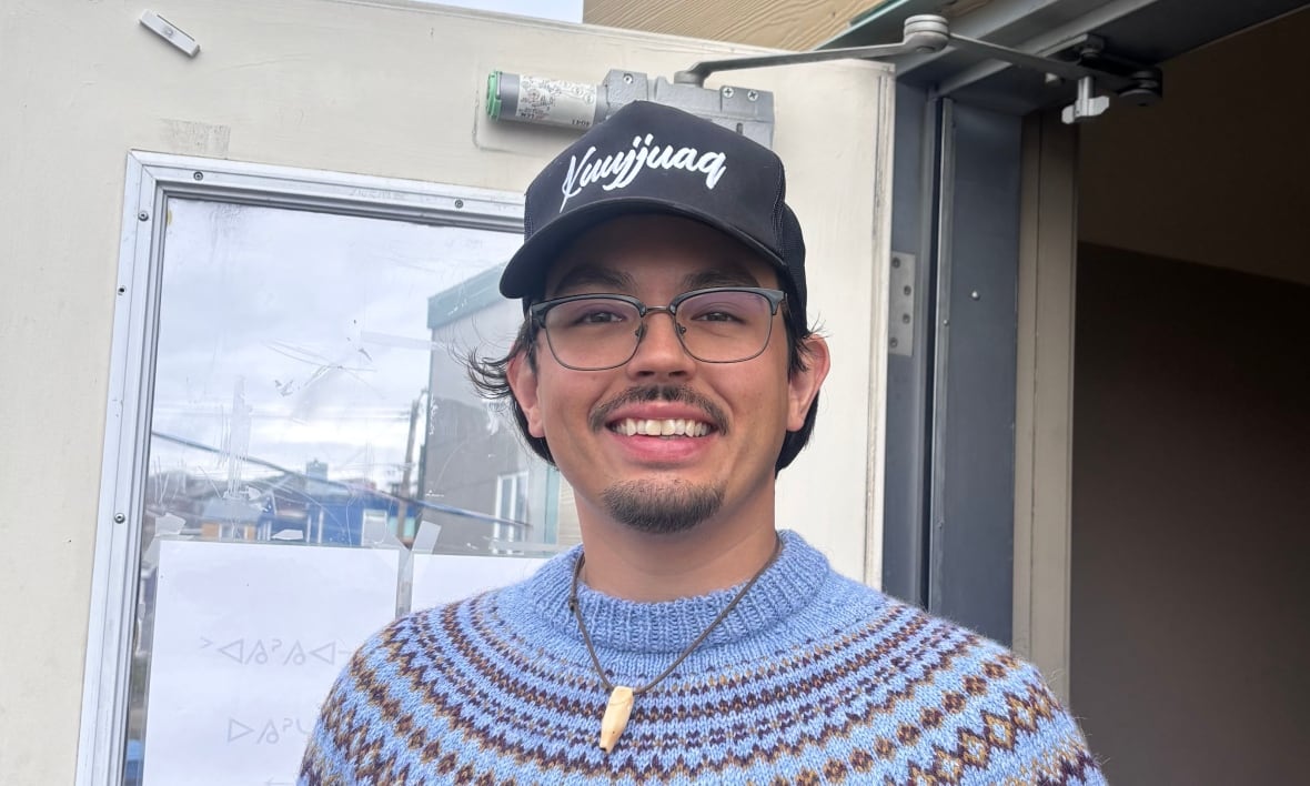 Man in blue sweater and baseball cap stands smiling in front of community centre.