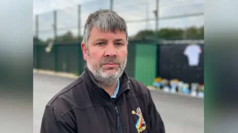 George Carden/BBC James Wilson pictured at the Chichester City FC football ground. James has black/grey hair and is wearing a black zip up jacket and blue shirt. The floral and card tributes can be seen in the background, though are partially blurred out.