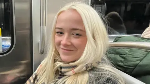 Cat Edge A woman sitting on a train and smiling at the camera. She has long blonde hair and is wearing a tartan patterned scarf. Behind her are metal train doors and a darkened window.