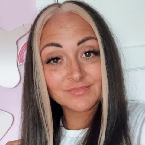 Charmaine Hines @mj_mum_of_3 A selfie of a woman with brown hair with blonde streaks, stood in front of a white and pink wall indoors