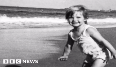 A black and white photograph showing three-year-old Cheryl Grimmer standing on a beach with waves gently lapping in the background. She has short fair hair and is wearing a white bathing costume.
