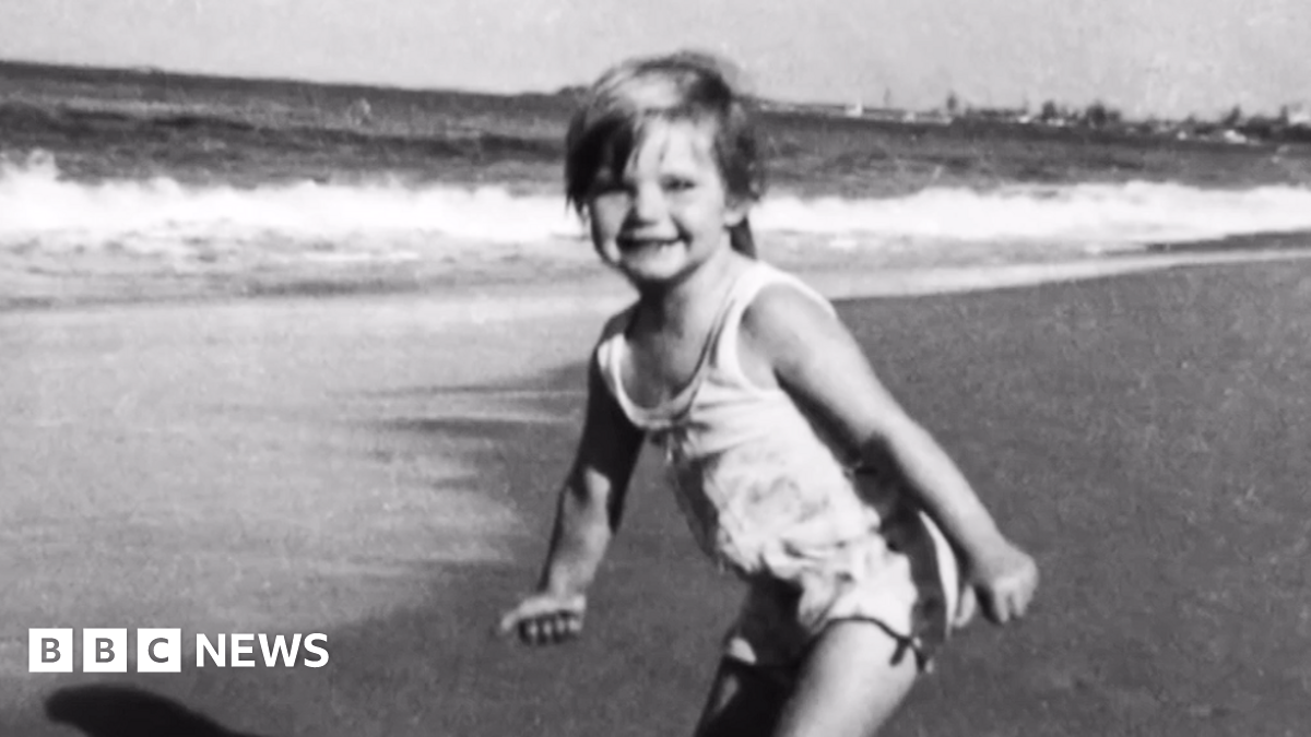 A black and white photograph showing three-year-old Cheryl Grimmer standing on a beach with waves gently lapping in the background. She has short fair hair and is wearing a white bathing costume.