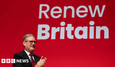 Sir Keir Starmer stands in front of a giant red screen with the slogan Renew Britain. He is wearing a dark suit with a white shirt and a red tie. He is applauding Chancellor Rachel Reeves, not shown.
