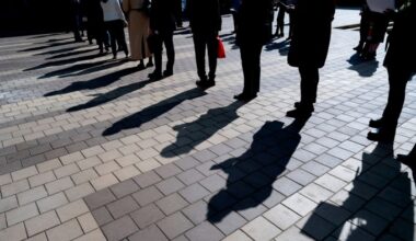 People stand in line casting long shadows on pavement as they wait to enter a job fair event.
