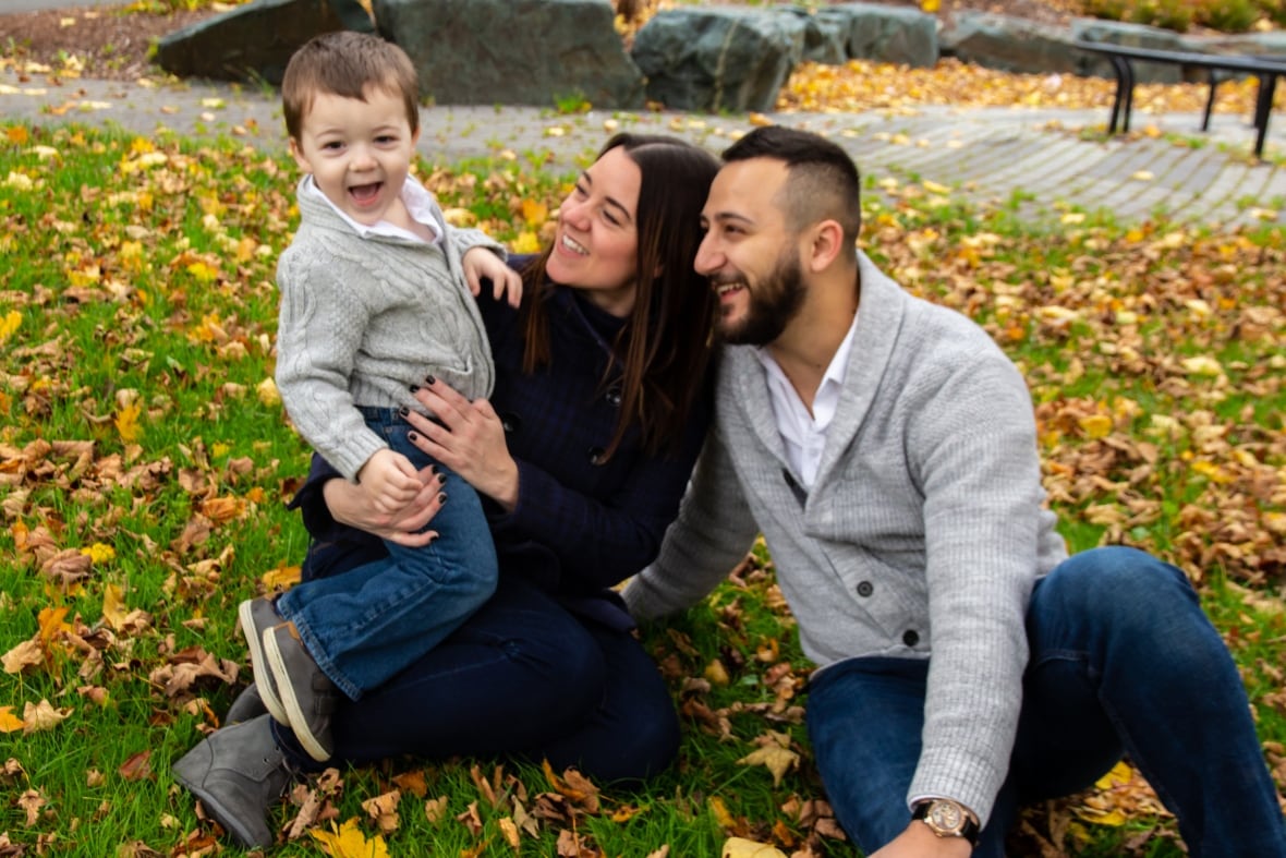 A man and woman sit in the grass of a park with fallen autumn leaves around them, smiling at a young boy in the woman's arms.