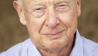 An older gentleman with white hair is seen wearing a blue and white striped button-up shirt, set against a blurred neutral brown backdrop.