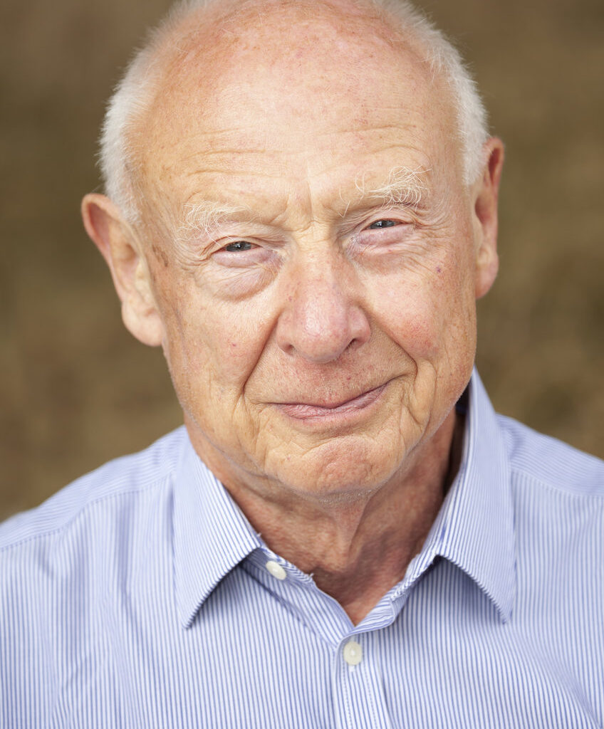 An older gentleman with white hair is seen wearing a blue and white striped button-up shirt, set against a blurred neutral brown backdrop.