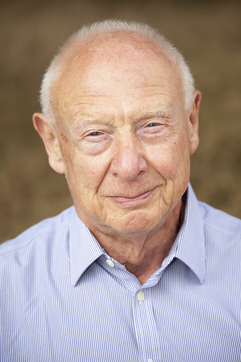 An older gentleman with white hair is seen wearing a blue and white striped button-up shirt, set against a blurred neutral brown backdrop.