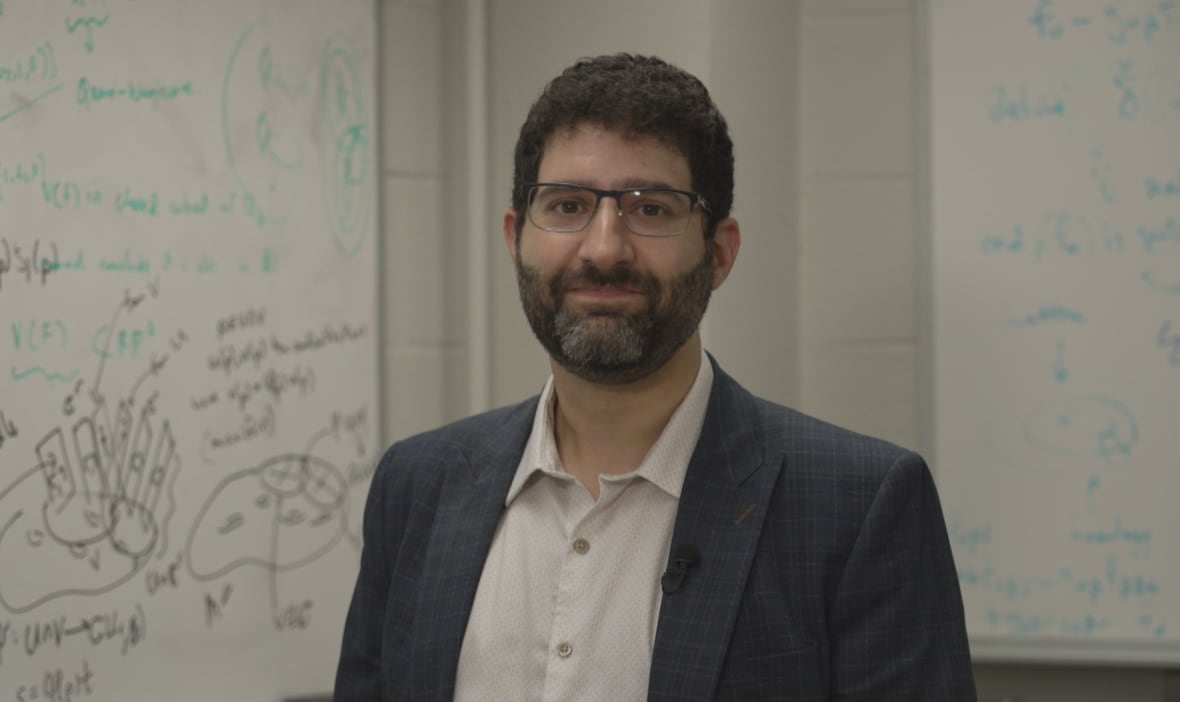 A man wearing glasses and a suit poses in front of a white board. 