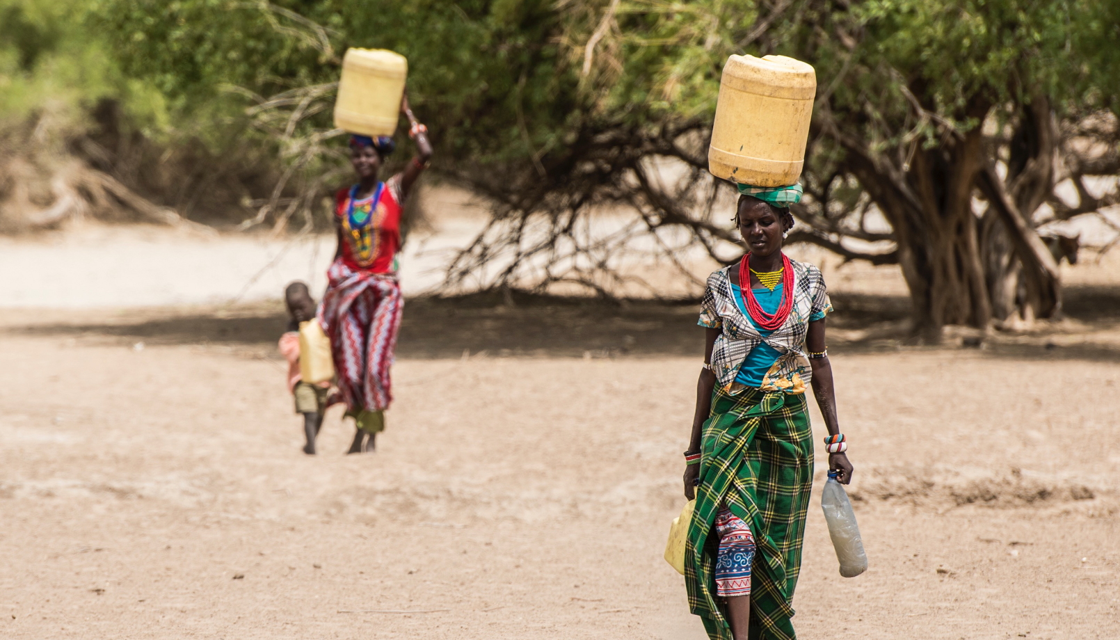 two women in colorful clothes walking through desert scrub, each carrying a plastic jerrycan on her head