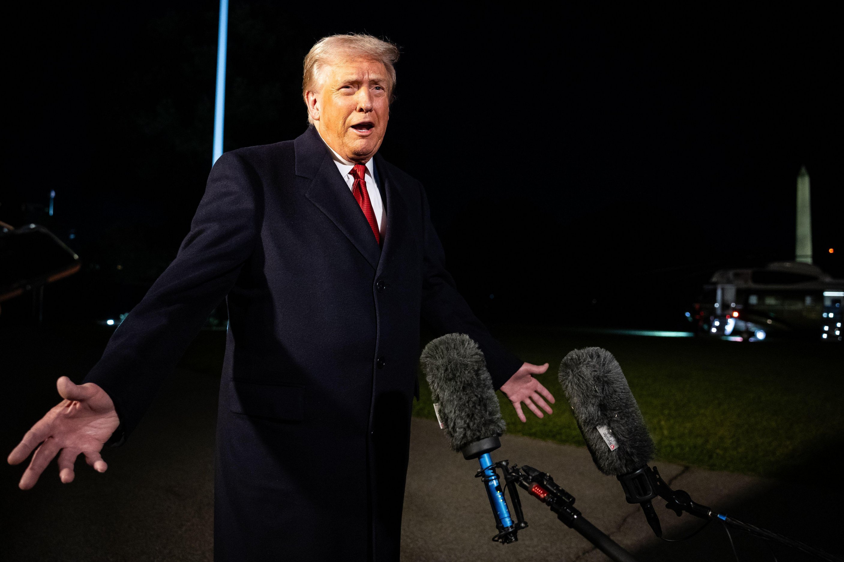 Donald Trump speaking to reporters at night with microphones in front of him and Marine One and the Washington Monument in the background.