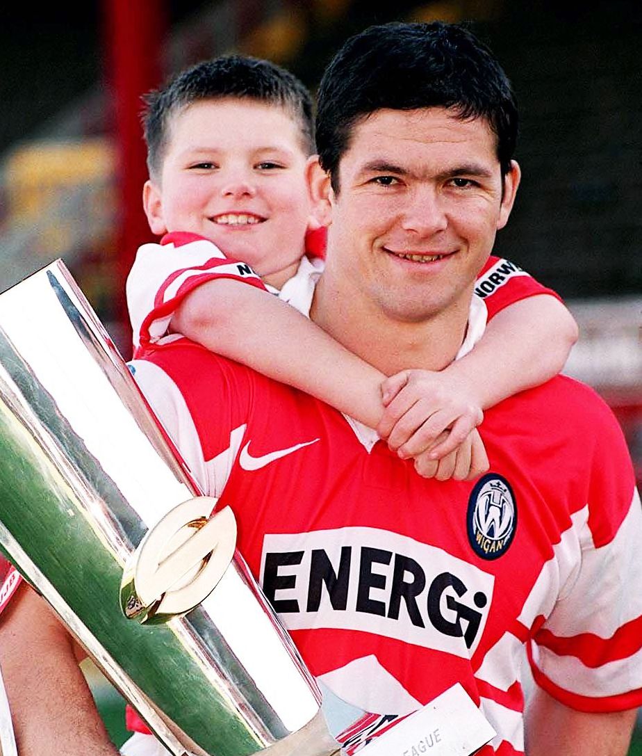 Andy Farrell holding a trophy with young Owen Farrell on his back, both smiling and wearing red and white jerseys.