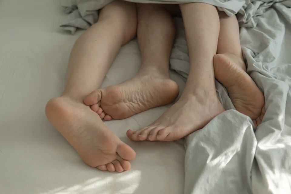 Three pairs of bare feet entangled under a light bedsheet, suggesting intimacy and relaxation