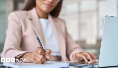 A woman, whose face is only half visible and slightly out of focus, wearing a pale  pink jacket, writes using a smart ballpoint pen on a spiral bound notebook. Her other hand is using the keyboard of a laptop that is open on the table in front of her.