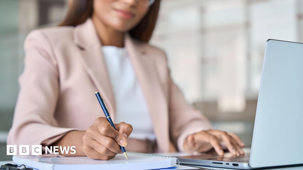 A woman, whose face is only half visible and slightly out of focus, wearing a pale  pink jacket, writes using a smart ballpoint pen on a spiral bound notebook. Her other hand is using the keyboard of a laptop that is open on the table in front of her.