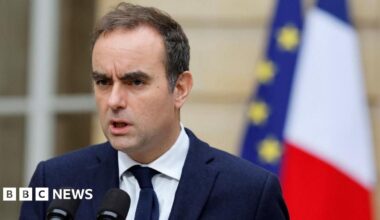 Sébastien Lecornu stands outside in front of two flags, French and EU, as he delivers a stern message in front of two microphones wearing a blue jacket and tie