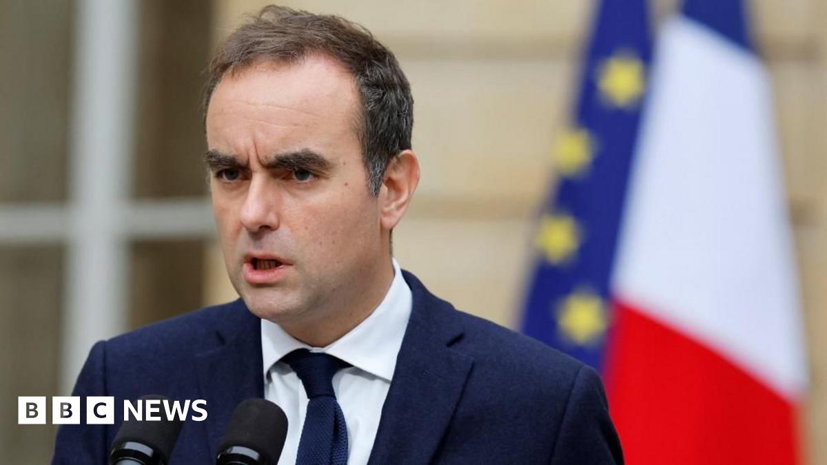 Sébastien Lecornu stands outside in front of two flags, French and EU, as he delivers a stern message in front of two microphones wearing a blue jacket and tie