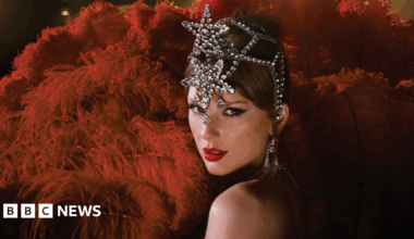 Taylor Swift is dressed as a showgirl with an elaborate, rhinestone headpiece. She is surrounded by crimson red feathers.