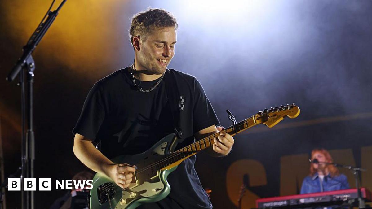 Sam Fender performs onstage for day two of the 2025 Pilgrimage Music & Cultural Festival at The Park at Harlinsdale Farm. He is playing a green electric guitar and is smiling down at his instrument. He is wearing a black T-shirt and silver chain. A keyboard player is behind him on stage.