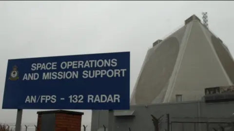 The exterior of the air force base. It's a tall, grey structure surrounded by small huts and barbed-wire.