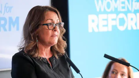 Getty Images Linden Kemkaran wearing glasses and talking into two podium microphones. Light blue and white backgrounds can be seen behind her.