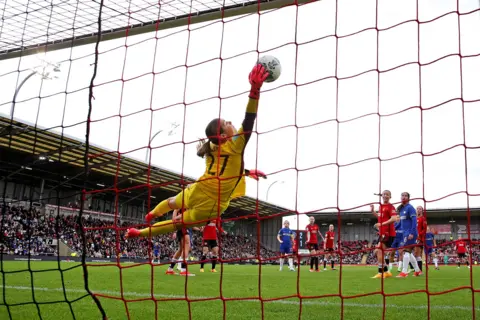The FA via Getty Images Mary Earps dives and saves the ball after Lauren James of Chelsea heads it towards the goal, she's diving in mid-air with a glove on the ball, while the photo is taken from the back of the goal showing the net in the foreground, and both Chelsea and Man Utd players in the background against the backdrop of the stadium and a grey sky, at Leigh Sports Village in April 2024.