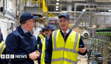 Peter Kyle being led around a glass factor in Cheshire - he is gesturing with his left hand, while standing among the machinery - he is wearing a baseball cap and a high visibility jacket
