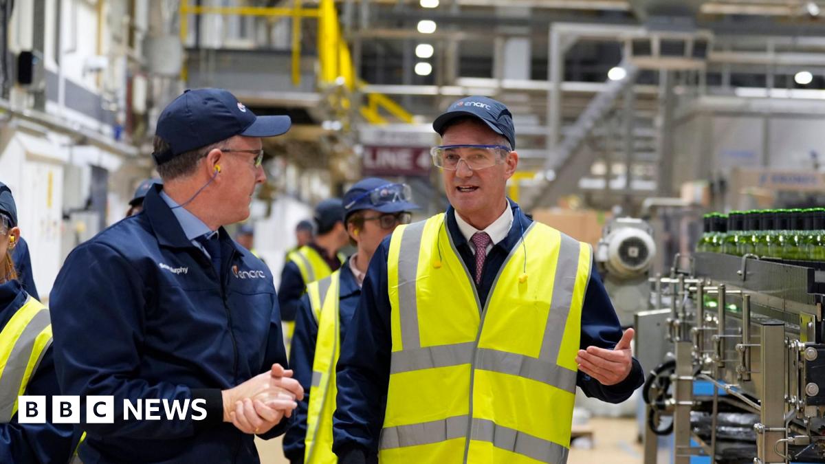 Peter Kyle being led around a glass factor in Cheshire - he is gesturing with his left hand, while standing among the machinery - he is wearing a baseball cap and a high visibility jacket