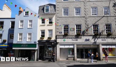 A photo of the middle of Guernsey's High Street. In the foreground are people standing waiting, while the background is a large grey building (Boots), while next to it is a tall skinning cream building with a a blue awning, alongside a blue building with a green sign for HS2.