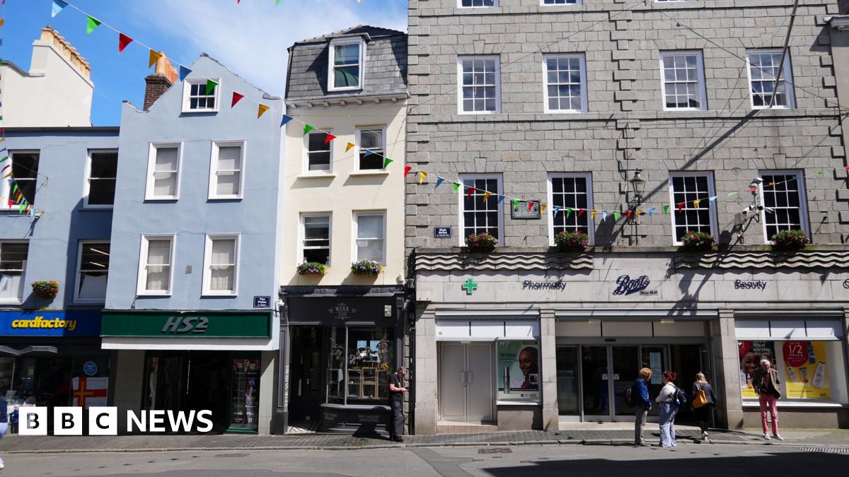 A photo of the middle of Guernsey's High Street. In the foreground are people standing waiting, while the background is a large grey building (Boots), while next to it is a tall skinning cream building with a a blue awning, alongside a blue building with a green sign for HS2.