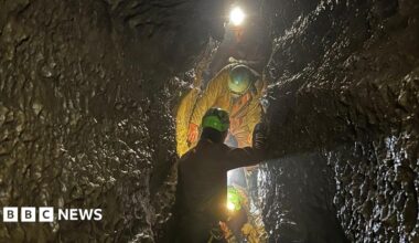 A man wearing a red jumpsuit and orange helmet with a torch attached hangs on ropes, next to a sheep. They are both in a cave.