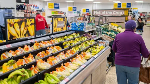 Getty Images A shopper in a purple sweater pushes a trolley in a Tesco supermarket beside a fresh fruit stand