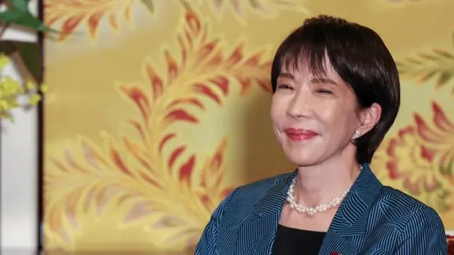 Takaichi smiling with her mouth closed. She is wearing a blue blazer and a white pearl necklace