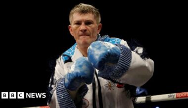 Ricky Hatton stands in a boxing ring. He is wearing a white tasselled suit and blue boxing gloves and punching the air.