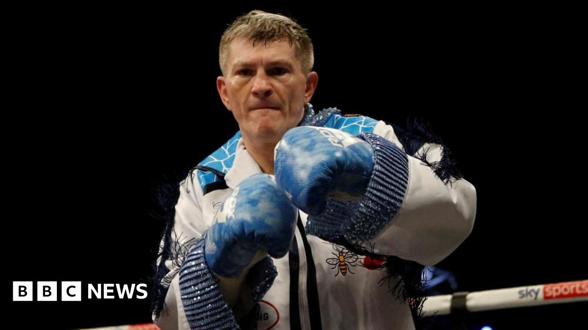 Ricky Hatton stands in a boxing ring. He is wearing a white tasselled suit and blue boxing gloves and punching the air.