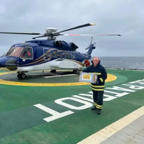 Sam Crowe Offshore worker holding a 'live bird' box on an offshore platform next to a helicopter, with word 'platform' is written in bold white letters on the deck.