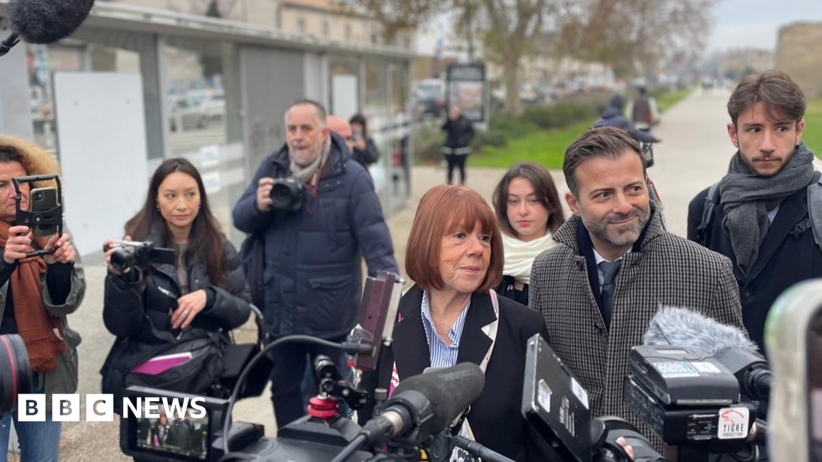Gisèle Pelicot is surrounding by members of the press carrying cameras. She is walking with her head held high with a faint smile. She has a red bob and is wearing a black coat. She is flanked by her legal team.