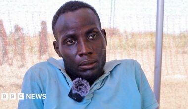 A head and shoulders shot of man in a light blue shirt. He has a clip mic attached to a collar.