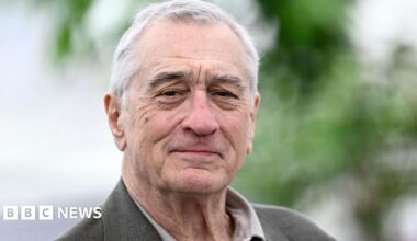 Robert De Niro, pictured from the shoulder up, with a neutral expression, wearing a lighjt grey suit jacket and open collar shirt.