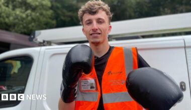 A young man stands next to a white van dressed in an orange jacket and black boxing gloves.