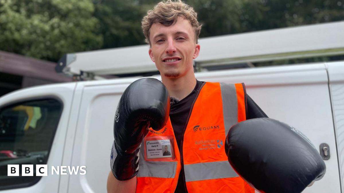 A young man stands next to a white van dressed in an orange jacket and black boxing gloves.