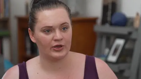 BBC A woman wears a purple tank top. Her hair is tied up in a bun.
