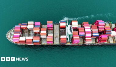 A large cargo ship staked with pink, blue and white container ships sails from right to left through the picture, a small trail of white foam in the picture.