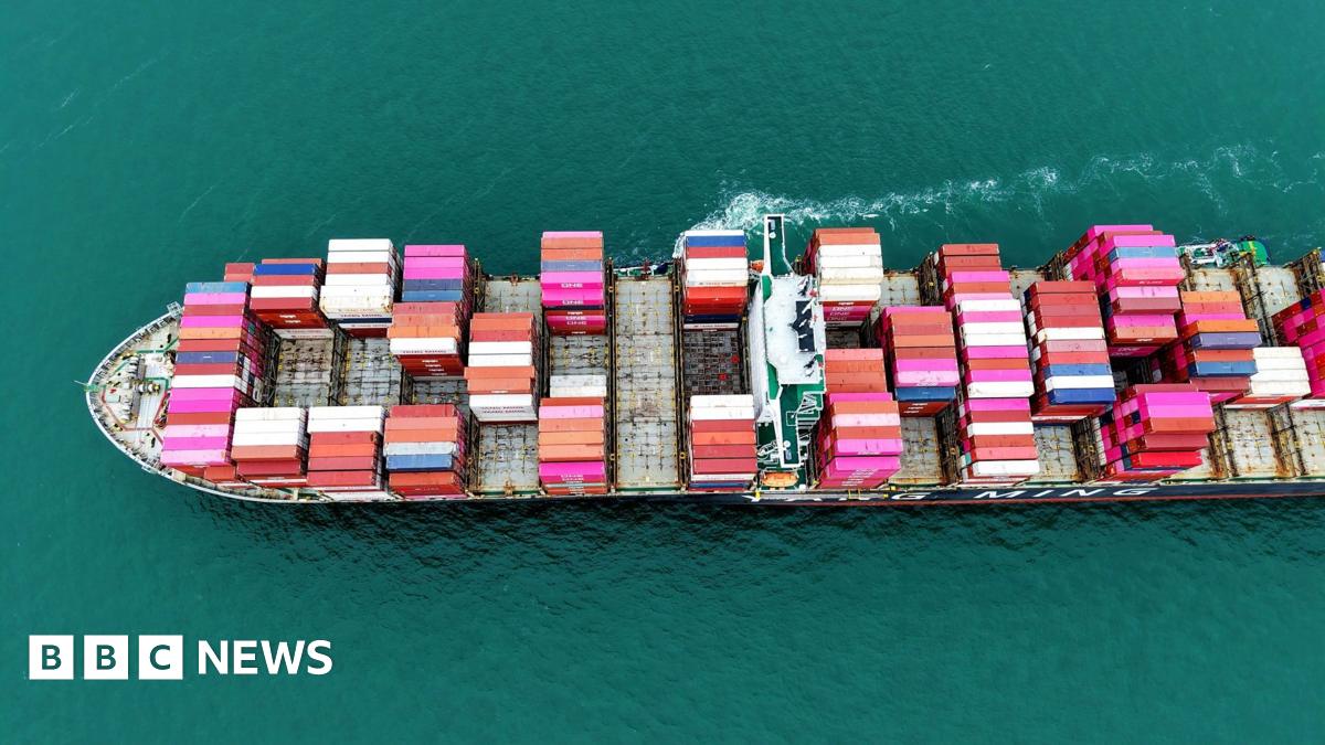 A large cargo ship staked with pink, blue and white container ships sails from right to left through the picture, a small trail of white foam in the picture.