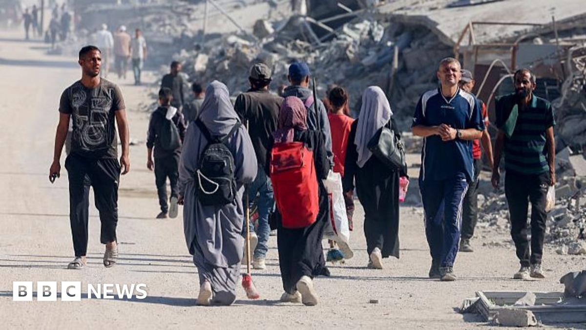 Various people walking along in Gaza, mainly away from the camera, with huge piles of debris in the background.