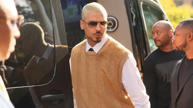 Brown, with bleached hair, sunglasses, and goatee and wearing a tan sweater vest over a white shirt, steps out of a ban as two men wearing black look on