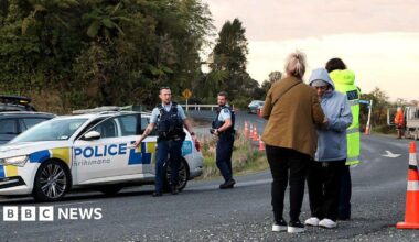 Two policemen standing near a police car, while two women wearing jackets and long pants stand on the side of the road. In the background is a tall green forest.